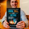 Business man sits smiling holding a book by titled Grow, Sell and Retire.